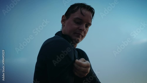 Surfer with wet hair puts on a wetsuit as the sun sets. He is on the beach and about to go in the water.
