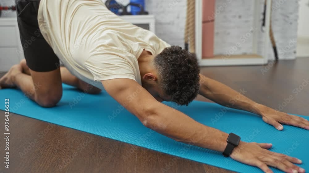 Young hispanic man doing a stretching exercise on a blue yoga mat in a gym with sports equipment in the background. Young hispanic man doing a stretching exercise on a blue yoga mat in a gym with sports equipment in the background.