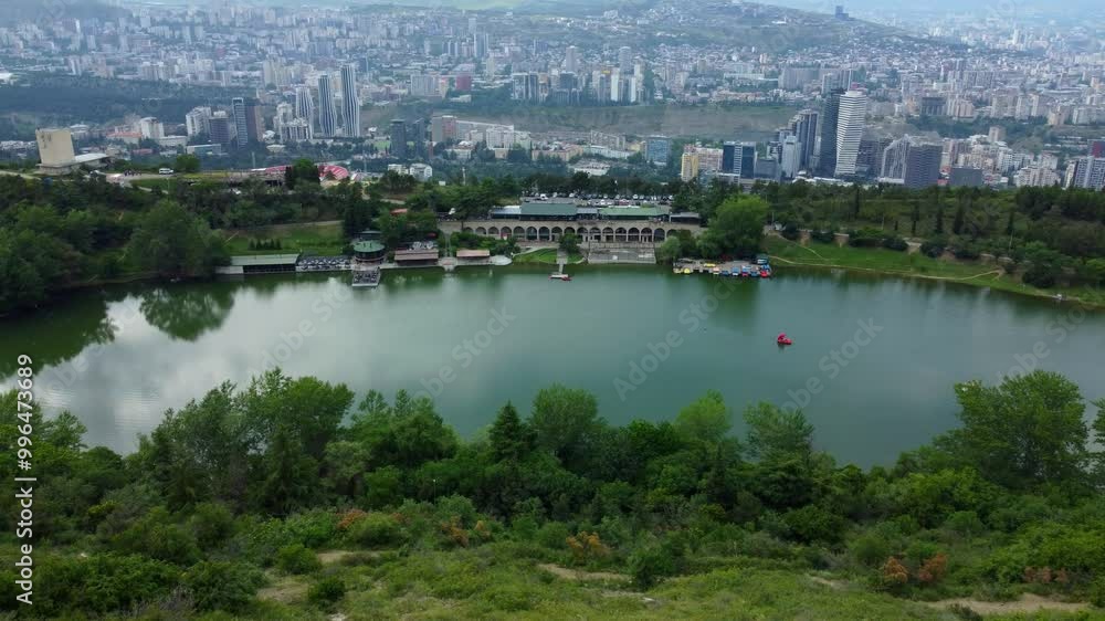 Clouds reflected in lake surrounded by forest. In background you can see city of Tbilisi with buildings and skyscrapers. Drone view of Turtle Lake.