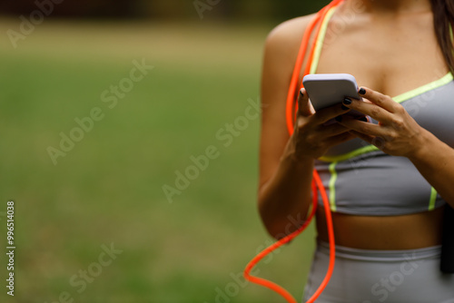 A girl who finished her workout in the park is sending messages on her phone, relaxing after physical activity, with nature in the background.