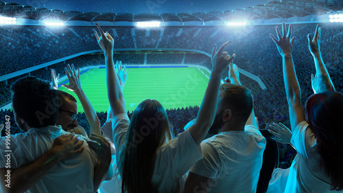 Canvas-taulu Fans in white shirts celebrate and pointing fingers toward soccer field, with field brightly illuminated and stadium packed with spectators