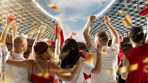 Fotografie Enthusiastic crowd in stadium raising their hands and red cups as golden confetti rains down during daytime match