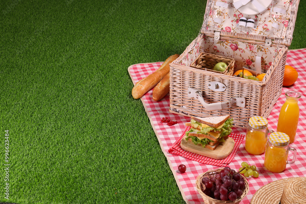 Fototapeta premium Picnic basket with tableware, different snacks, juice and hat on green grass outdoors, space for text