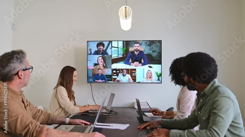 A hybrid team meeting where in-person attendees discuss and collaborate with remote colleagues through a video conference displayed on a wall-mounted screen. Concept of connectivity and flexibility
