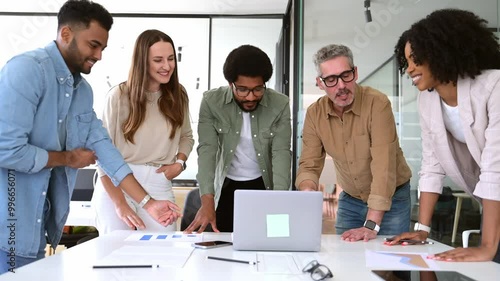 Diverse group of professionals leans in towards a laptop, sharing a moment of collaboration and interaction in their modern workspace. The energy and engagement of a diverse team working on a project