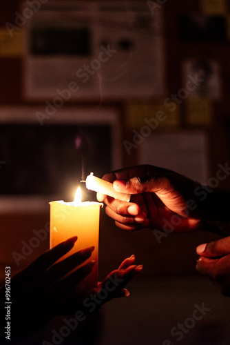 Close-up shot of hands lighting a candle in a dimly lit room, with a bulletin board and notes visible in the background, creating a mysterious and contemplative atmosphere.