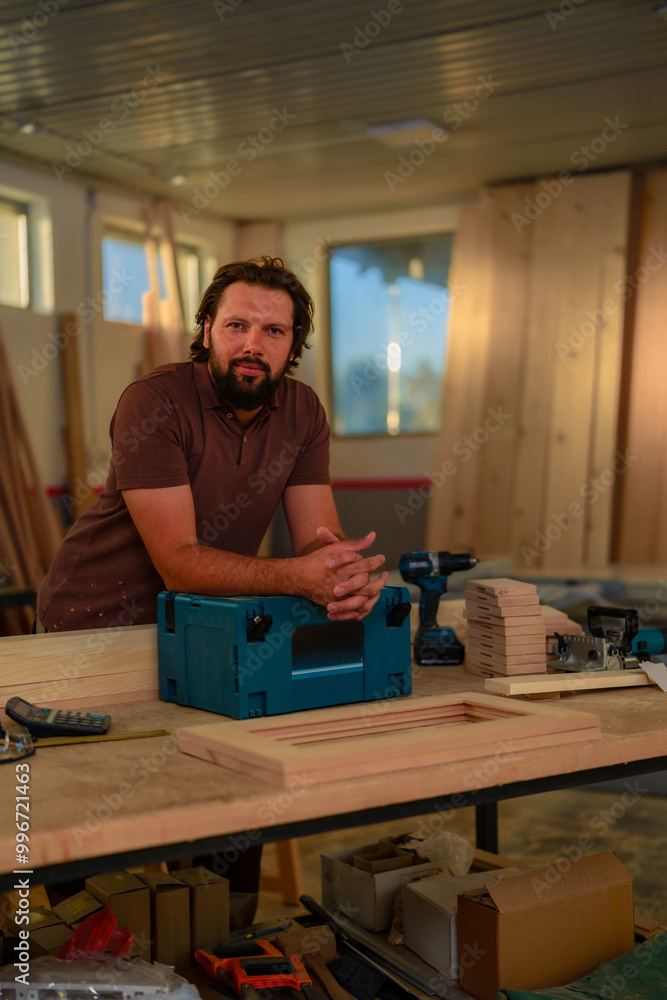Man with a beard stands confidently in a woodworking workshop, leaning on a blue tool box. The workspace is filled with wooden planks, tools, and equipment. Man with a beard stands confidently in a woodworking workshop, leaning on a blue tool box. The workspace is filled with wooden planks, tools, and equipment.