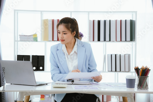 Confident Asian businesswoman in modern office smiling while working on laptop, analyzing reports, planning marketing strategies and leading with energy and positivity.