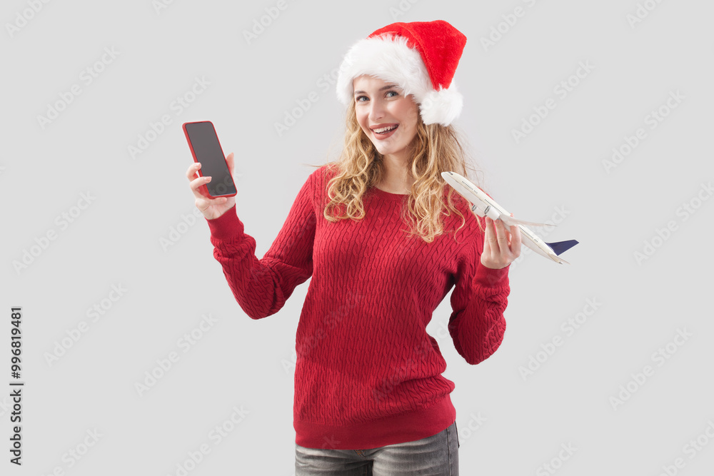 Happy merry Christmas woman wearing Santa hat holds cellphone and showing model airplane. Concept for Travel booking online, advertising banner and poster for winter holiday, isolated background Happy merry Christmas woman wearing Santa hat holds cellphone and showing model airplane. Concept for Travel booking online, advertising banner and poster for winter holiday, isolated background