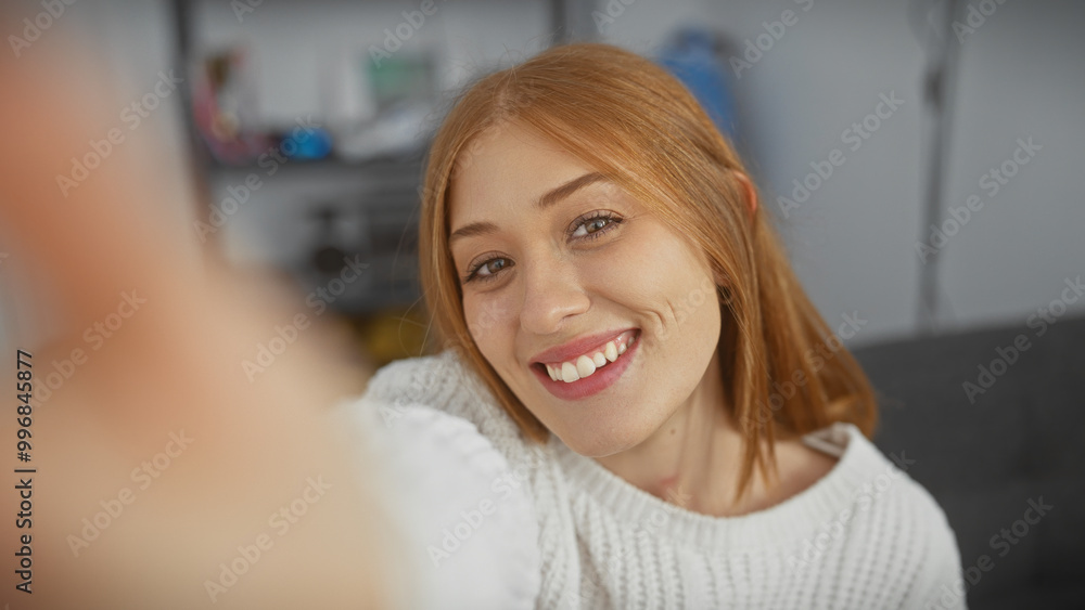 A young, smiling caucasian woman with redhead hair in a cozy indoor setting poses for a casual portrait at home. A young, smiling caucasian woman with redhead hair in a cozy indoor setting poses for a casual portrait at home.