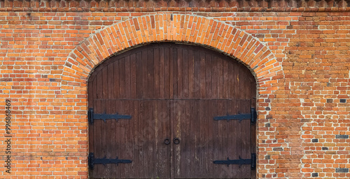 Fotografie Old brick wall with arch and closed wooden gate with old-style door hinges and r