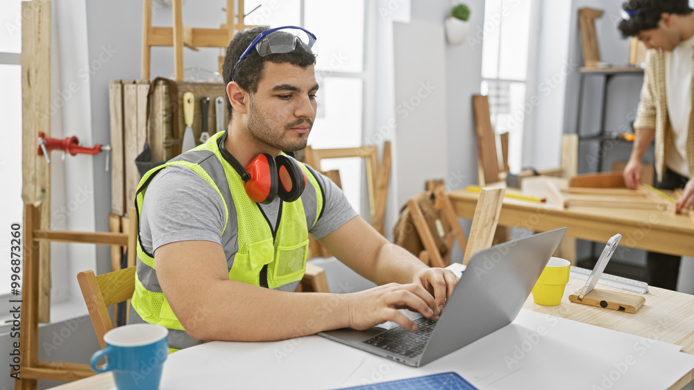 Two men work in a bright carpentry workshop, one using a laptop while the other measures wooden planks.