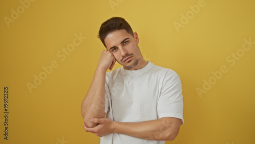 A pensive young hispanic man with a neat beard poses against a plain yellow background, exuding casual confidence.