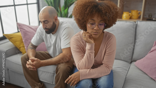 Φωτογραφία A thoughtful interracial couple sitting apart on a grey sofa, displaying tension and contemplation in a modern apartment