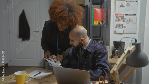 A woman and a man, possibly detectives, examine documents in a well-equipped investigation office. photo