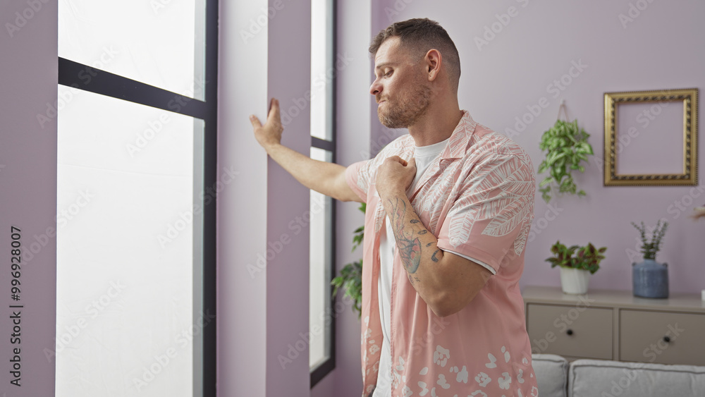 Young tattooed man in a modern apartment touching windowpane gently, reflecting calm and contemplation. Young tattooed man in a modern apartment touching windowpane gently, reflecting calm and contemplation.