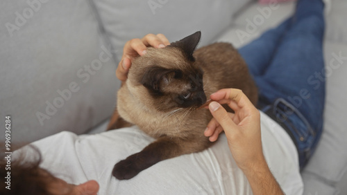 Young hispanic man bonding with his siamese cat on a couch at home