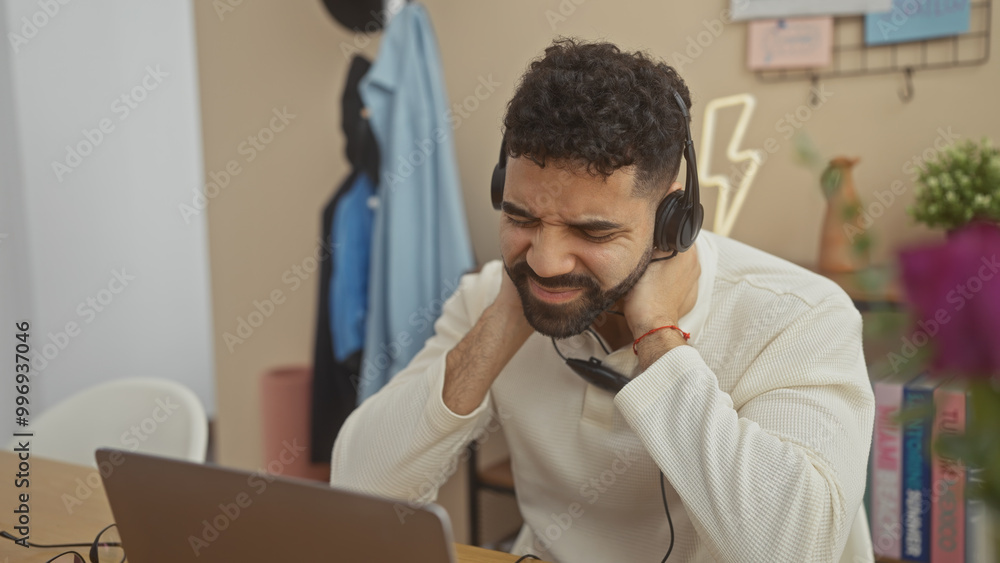 Hispanic man with beard experiencing neck pain at home in front of laptop with headphones. Hispanic man with beard experiencing neck pain at home in front of laptop with headphones.
