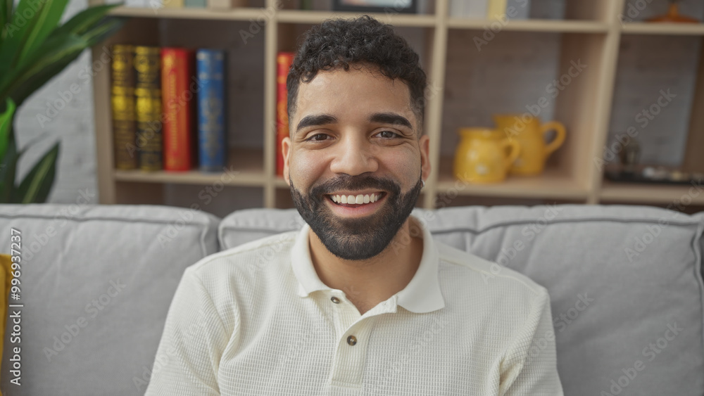 Smiling bearded man sitting in a modern living room with bookshelf and cozy couch, portraying casual home lifestyle. Smiling bearded man sitting in a modern living room with bookshelf and cozy couch, portraying casual home lifestyle.