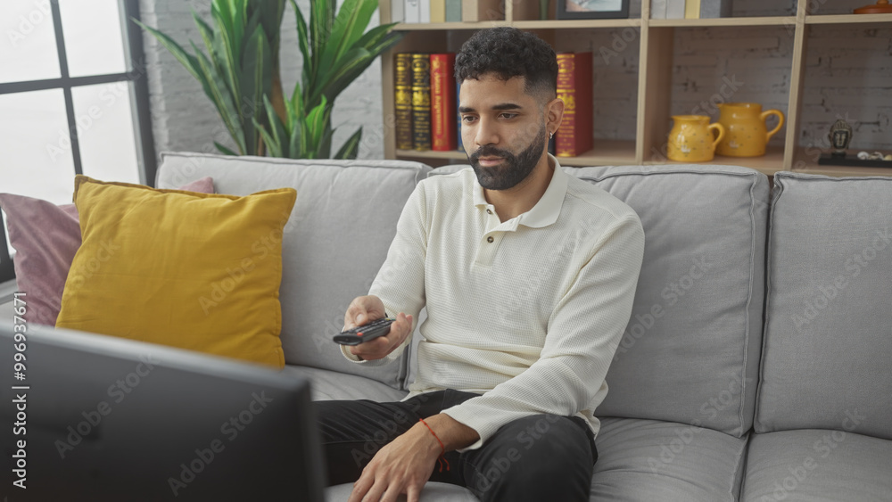 A young man with a beard watching television in a modern living room, holding a remote control A young man with a beard watching television in a modern living room, holding a remote control