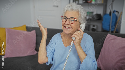 Bild auf Leinwand A cheerful senior woman in glasses speaking on a landline phone in a cozy living room, expressing happiness and conversation