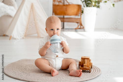 a baby drinks water from a bottle or a drinking cup sitting on the floor in a bright bedroom, a smiling, joyful baby boy in a white bodysuit eats or drinks photo