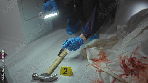 Detective examines a bloody hammer at a numbered indoor crime scene with sheets photo