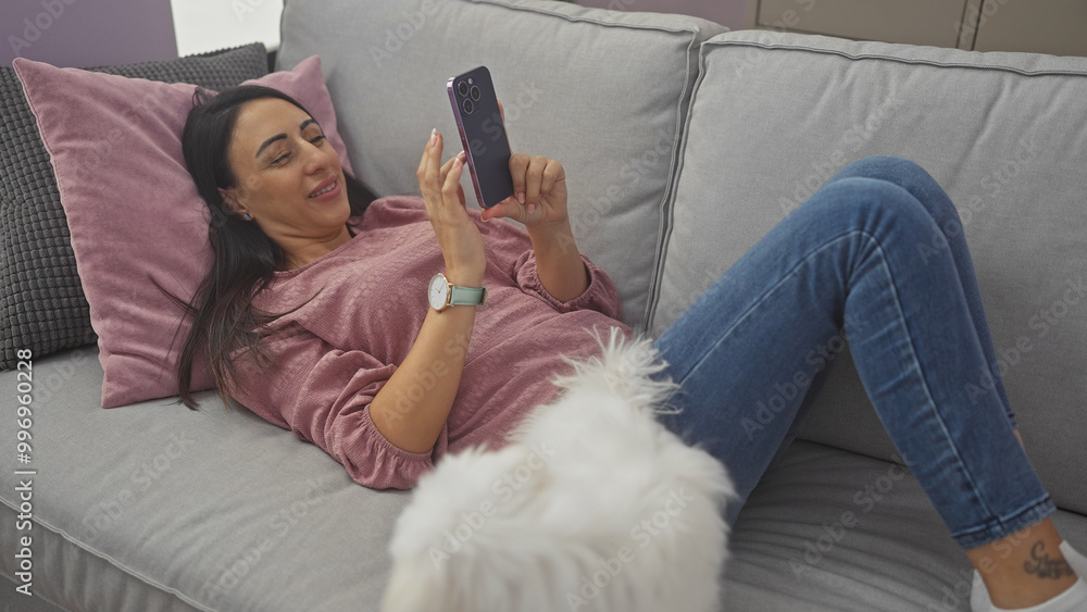 A smiling young woman relaxes with her smartphone on a sofa next to her bichon maltese in a cozy living room. A smiling young woman relaxes with her smartphone on a sofa next to her bichon maltese in a cozy living room.