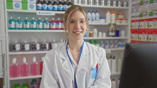 Smiling young woman pharmacist in white lab coat standing in a pharmacy with shelves of products