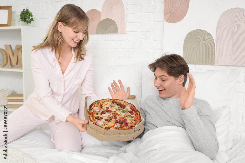 Young woman holding cardboard box with tasty pizza and her surprised boyfriend sitting on bed in bedroom Young woman holding cardboard box with tasty pizza and her surprised boyfriend sitting on bed in bedroom