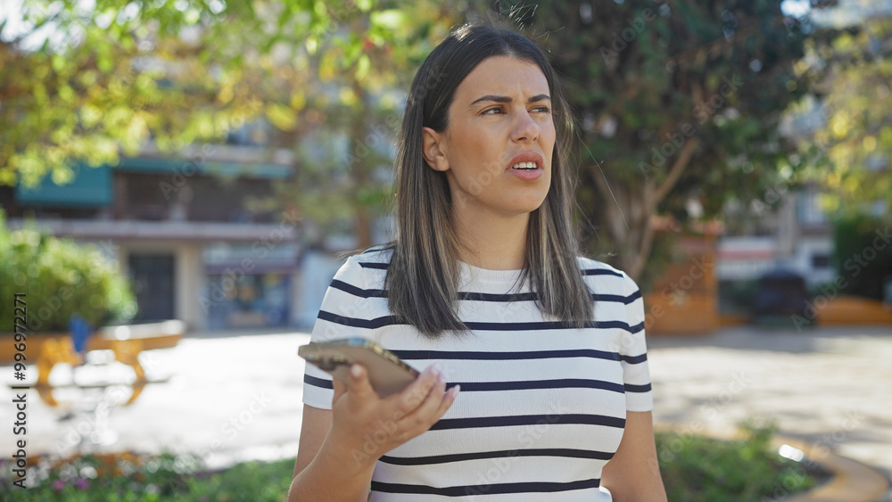A puzzled young hispanic woman holding a smartphone in an urban park setting. A puzzled young hispanic woman holding a smartphone in an urban park setting.