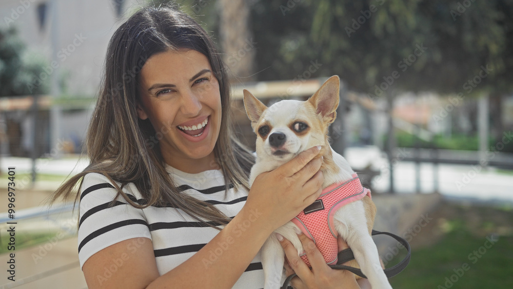 A young, beautiful hispanic woman is smiling while holding her chihuahua dog in a lively urban park setting. A young, beautiful hispanic woman is smiling while holding her chihuahua dog in a lively urban park setting.