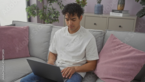 Hispanic man using laptop on couch in a modern living room setting.