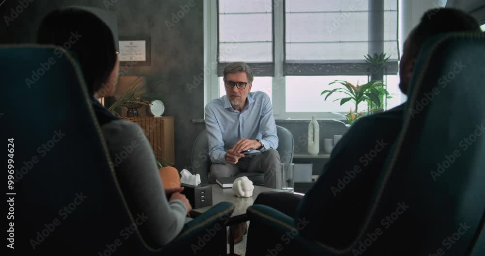 Mature psychotherapist talks to young family, provides couples therapy. Back view of multiethnic couple sitting on chairs, listening psychologist on counseling session. Psychology and sexology concept