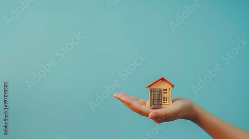 A hand holding a small model house against a blue background, symbolizing home ownership or real estate.