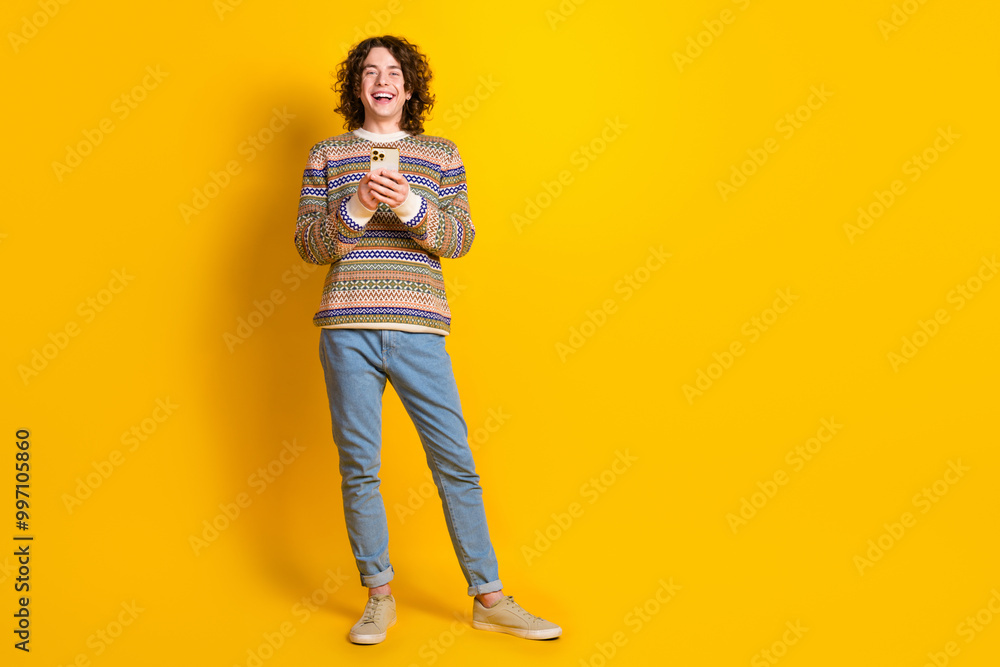 Full size photo of optimistic guy dressed ornament sweater holding smartphone read notification isolated on yellow color background Full size photo of optimistic guy dressed ornament sweater holding smartphone read notification isolated on yellow color background