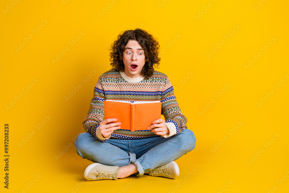 Full size photo of astonished guy dressed ornament sweater sitting on floor reading interesting book isolated on yellow color background Full size photo of astonished guy dressed ornament sweater sitting on floor reading interesting book isolated on yellow color background