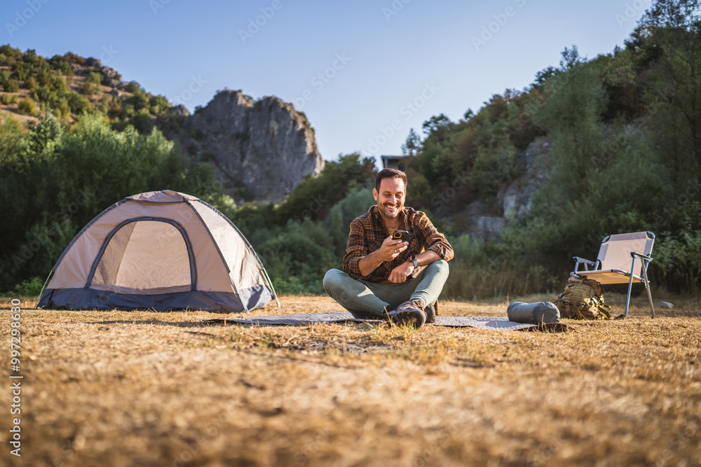 Fototapeta premium Adult man sit near tent and use mobile phone on camping trip