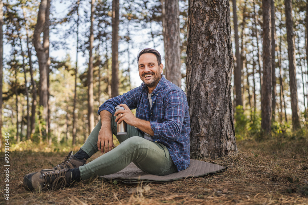 Adult man sit on mat in front tree and drink coffee from thermos