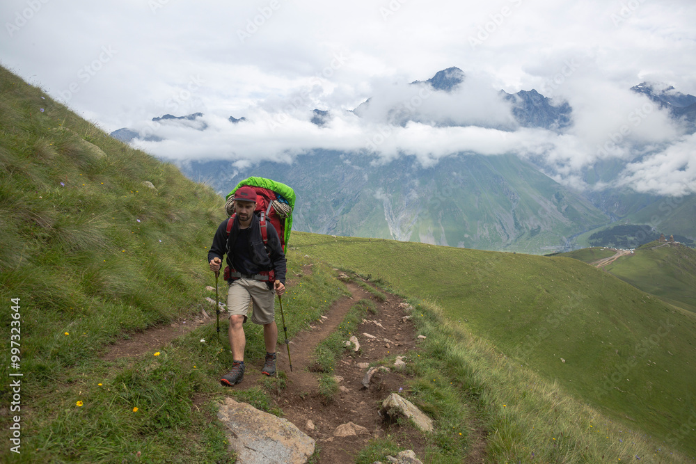 Obraz premium A hiker pauses on a mountain trail, carrying a large pack, with majestic peaks shrouded in mist behind him