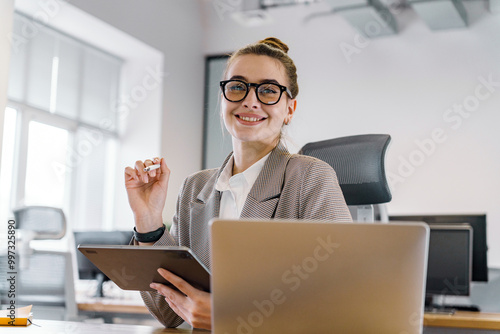 Professional Woman Smiling at Desk While Holding Tablet in Modern Office Environment During Daylight Hours photo