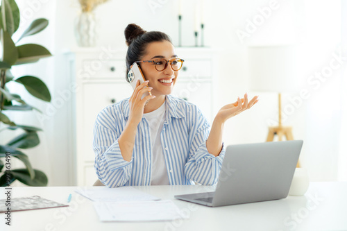 Mobile phone talk. Confident attractive indian or arabian business woman talking by smartphone about strategy with a client or partner while working day on her office with a laptop on the desk, smiles