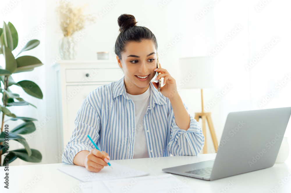 Smartphone conversation. Successful young beautiful caucasian woman with glasses, talking on mobile phone and using laptop while sitting at her working place, talk over idea with a client, smiling Smartphone conversation. Successful young beautiful caucasian woman with glasses, talking on mobile phone and using laptop while sitting at her working place, talk over idea with a client, smiling