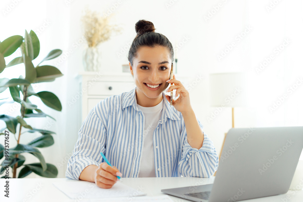 Phone talk concept. Successful caucasian business woman in blue shirt, talking on mobile phone and uses laptop while sitting at her workplace, discuss a project with client, smiles at camera friendly Phone talk concept. Successful caucasian business woman in blue shirt, talking on mobile phone and uses laptop while sitting at her workplace, discuss a project with client, smiles at camera friendly
