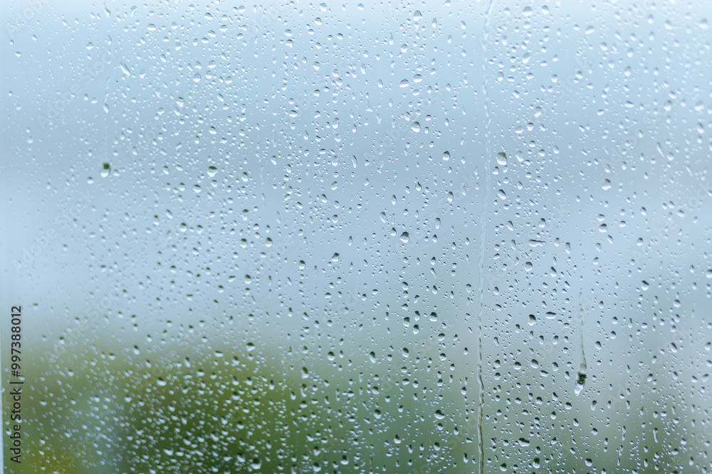 Rainy day with droplets on a windowpane, creating a blurred view of greenery outside during overcast weather Rainy day with droplets on a windowpane, creating a blurred view of greenery outside during overcast weather