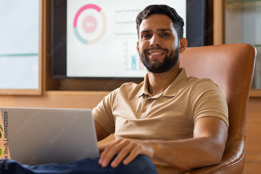 Smiling man using laptop in office with charts on screen in background Smiling man using laptop in office with charts on screen in background