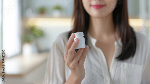 Woman holding small bottle of weight-loss pills, looking determined and focused in a minimalist, well-lit room with copy space for text photo