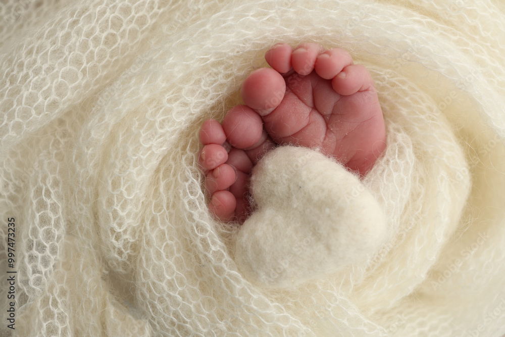 Obraz premium The tiny foot of a newborn baby. Soft feet of a new born in a white wool blanket. Close up of toes, heels and feet of a newborn. Knitted white heart in the legs of a baby. Macro photography. 