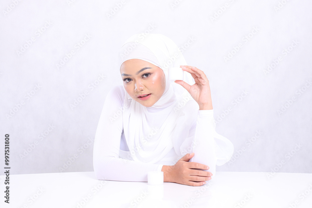 Beautiful young hijab woman expression looking down at camera hand holding cosmetic cream on left side of face for daily skin care routine needs on white background. Facial treatment, Perfect skin Beautiful young hijab woman expression looking down at camera hand holding cosmetic cream on left side of face for daily skin care routine needs on white background. Facial treatment, Perfect skin