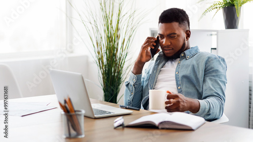 Multitasking concept. Millennial african american employee talking on cellphone, working on laptop and drinking coffee at workplace, copy space